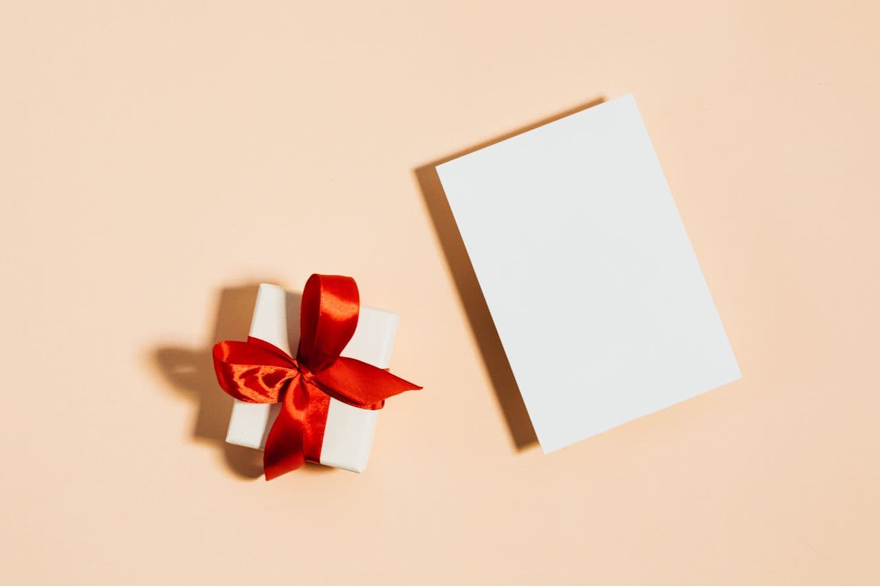 White gift box tied with a red ribbon beside a blank card on a peach background.